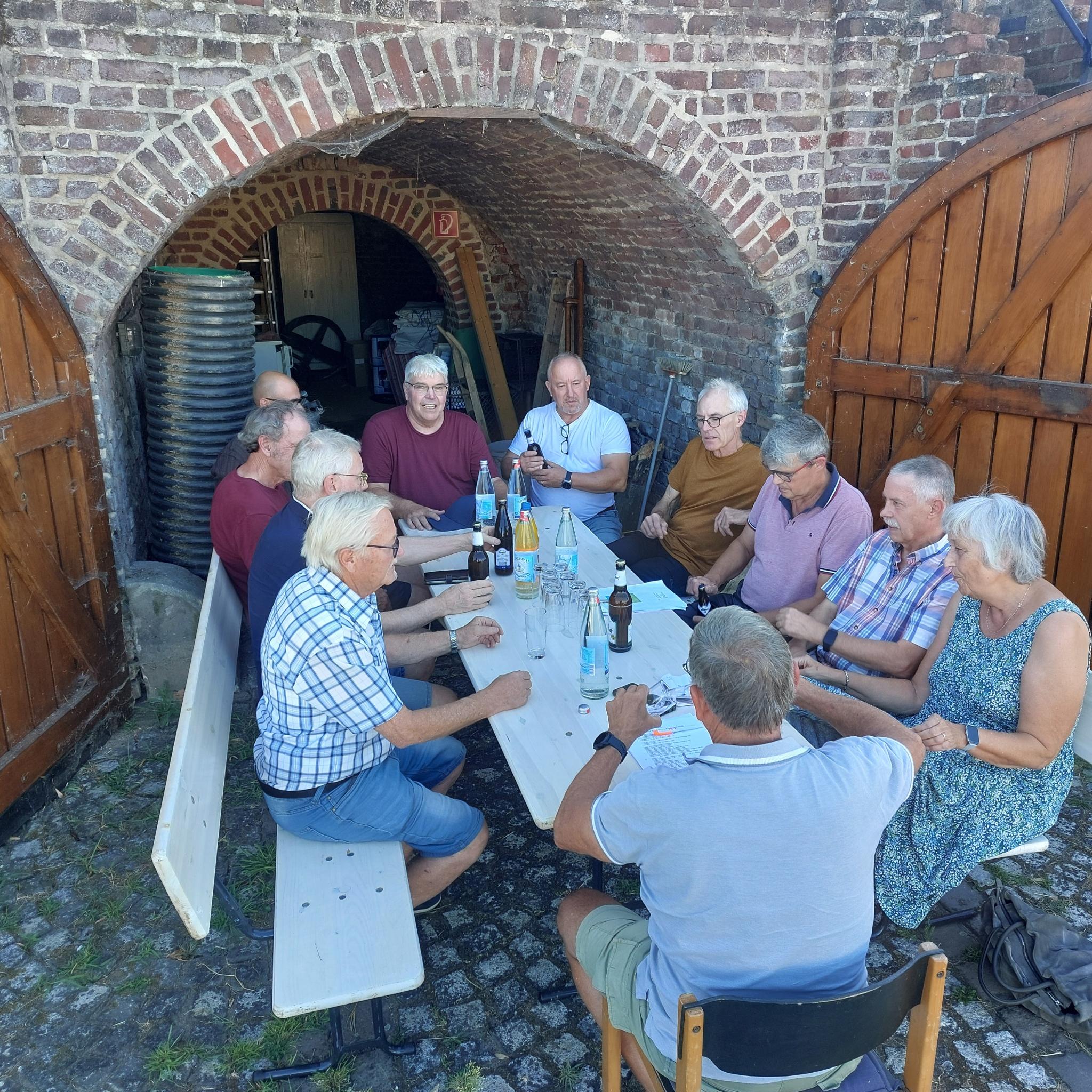Mehrere Menschen sitzen vor einem Tor, die Türen des Tores stehen offen. Die Menschen sitzen auf Bänken.