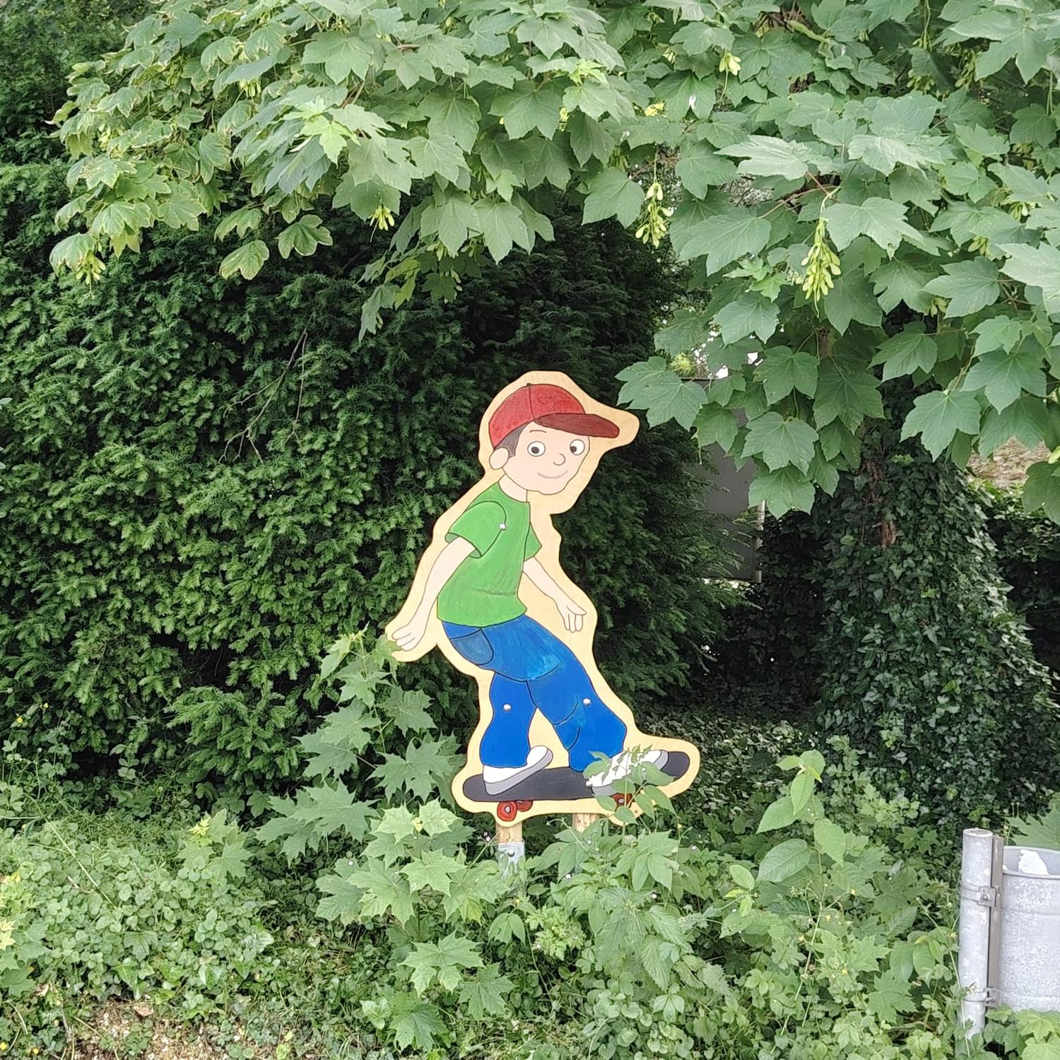 Im Grünen ist ein Junge auf einem Skateboard mit einer roten Käppi, grünem T-Shirt und einer blauen Hose.